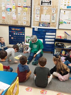 Pastor Nanny, masked, sitting on the floor in a preschool classroom, telling a story to the preschoolers.