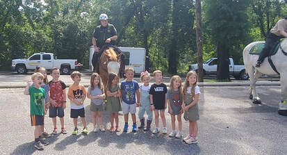A photo of 11 preschool children outside on a sunny day. They are in a parking lot and watching two officers from the Escambia County Mounted Posse, who are on horseback, as they visit the preschool.
