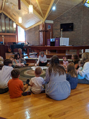A photo taken inside St. Paul Lutheran during worship. Children are seated, gathered around and facing a seated adult who is telling a story with manipulatives. Another adult is sitting behind the group, holding a guitar.
