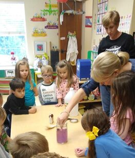 Two preschool teachers demonstrating a science experiment. Preschoolers surround the table and watch with curiosity.
