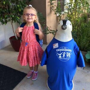 A young blonde preschool girl wearing glasses, a red dress, and a backpack. She has a huge smile and is standing at the entrance to St. Paul Preschool, next to a penguin statue. The penguin is wearing a blue St. Paul Preschool t-shirt.